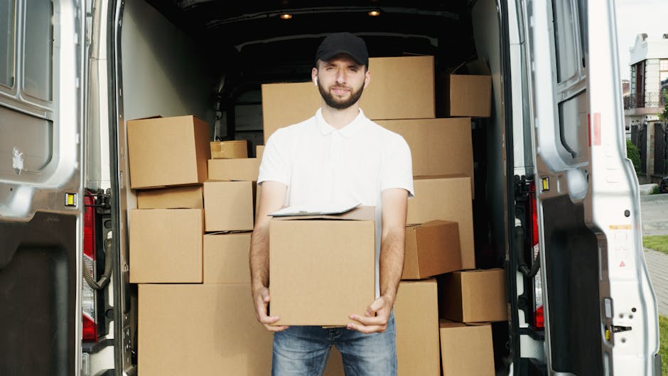 A man wearing a white shirt, black cap, and jeans stands inside an open moving van, holding a cardboard box with both hands. The van's interior is filled with neatly stacked cardboard boxes of various sizes, some placed directly on the floor and others on top of each other. The sides of the van are visible, with the rear doors open to the outside, where a paved pathway and residential buildings can be seen. The scene captures the loading process during a home relocation, with packing materials and boxes ready for transport. The lighting indicates daytime, with natural light illuminating the interior and exterior environment. Man With a Van Isleworth, a professional removals service, is involved in this furniture transport and packing and moving activity as part of their same-day delivery service in Isleworth.