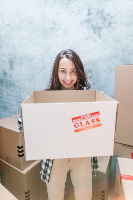 A person wearing a dark short-sleeved shirt with tattooed arms and a black wristwatch is carrying a large cardboard box on their shoulder during a home relocation process. The individual’s face is n