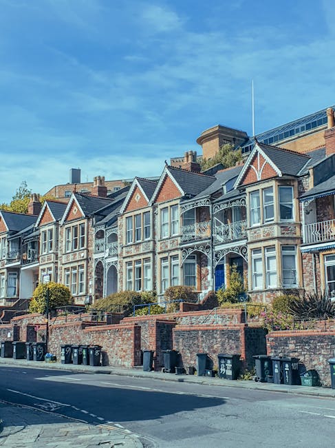 A row of Victorian-style terraced houses in Isleworth, TW7, featuring ornate ironwork balconies, bay windows, and brick facades with decorative trim. Several houses are set behind brick retaining walls with small front gardens, some with bushes and plants. The street in front of the houses has a paved sidewalk and black wheelie bins lined along the curb. Bright daylight illuminates the scene, with a clear blue sky overhead. The image, associated with house removals and relocation services, suggests a residential area where furniture transport and packing are typical activities for local movers like Man With a Van Isleworth. The scene is calm, well-maintained, and representative of typical housing stock in the area, ideal for a home relocation guide focused on TW7 streets.