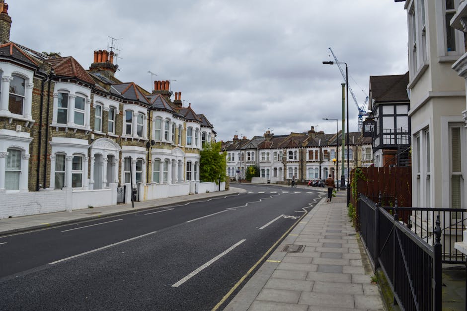 A deserted residential street in Isleworth, featuring a row of Victorian and Edwardian terraced houses with white and brick facades, bay windows, and red-tiled roofs. There are chimneys on the rooftops, and some houses have small front gardens enclosed by low walls or fences. The street is paved with asphalt and has designated parking lanes, with a few vehicles parked along the curb. The pavement is made of concrete slabs and runs parallel to a black metal railing fence on the right side of the image. A person wearing a brown jacket and dark trousers is walking away from the camera, while another person stands near parked cars further down the street. The sky is overcast with grey clouds, and there is a traffic crane visible in the background, indicating ongoing construction or building work. This scene reflects a typical environment where house removals or furniture transport might occur, with the street suitable for loading/unloading items during a home relocation, supported by local removals companies such as Man With a Van Isleworth.