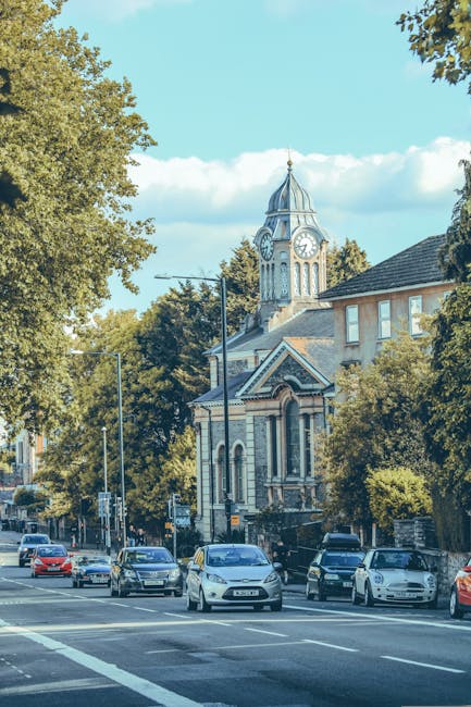 A row of Victorian-style terraced houses in Isleworth, TW7, featuring ornate ironwork balconies, bay windows, and brick facades with decorative trim. Several houses are set behind brick retaining walls with small front gardens, some with bushes and plants. The street in front of the houses has a paved sidewalk and black wheelie bins lined along the curb. Bright daylight illuminates the scene, with a clear blue sky overhead. The image, associated with house removals and relocation services, suggests a residential area where furniture transport and packing are typical activities for local movers like Man With a Van Isleworth. The scene is calm, well-maintained, and representative of typical housing stock in the area, ideal for a home relocation guide focused on TW7 streets.