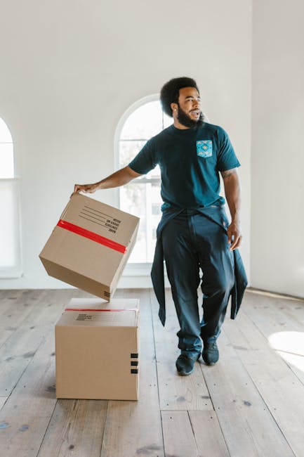 A person wearing a dark green shirt and navy blue trousers is holding and inspecting a large cardboard moving box inside a property. The box is sealed with packing tape and features a red and white ca