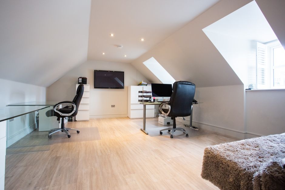 A spacious home office located in an attic room with angled ceilings and large skylight windows allowing natural light to illuminate the space. The room features light wood flooring, white walls, and 