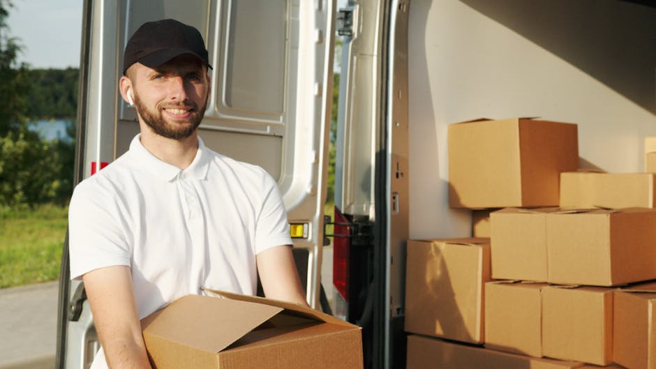 A man with a beard and wearing a black cap and a white polo shirt is standing outside near the open sliding door of a white van, preparing for a home relocation. He is holding a large cardboard box, w