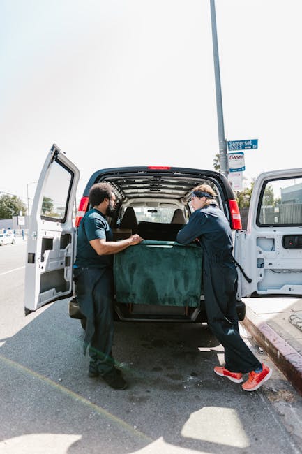 A man wearing a yellow jacket and jeans is loading a cardboard box into the back of a white Man With a Van Isleworth moving van parked on a residential street in Isleworth. The van's rear doors are op