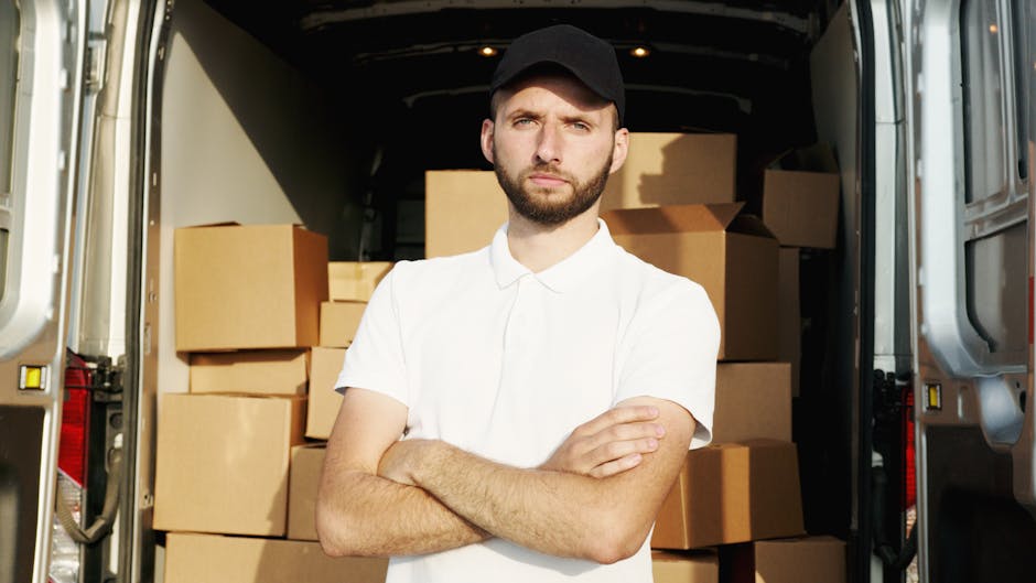 A man wearing a white t-shirt and jeans is standing on the roadside next to a white moving van with its rear doors open, preparing to load or unload cardboard boxes. The boxes vary in size and are sta